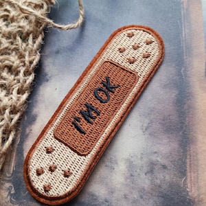 May include: An embroidered patch shaped like an adhesive bandage with a tan textured surface and a dark brown centre. The text I'M OK is stitched in black thread across the centre. The patch features a dark brown border and small brown accents.
