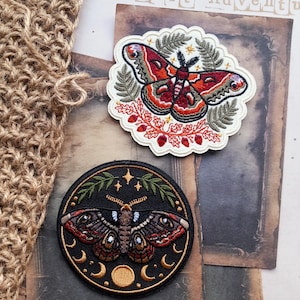 May include: Two embroidered patches featuring moth designs. The top patch is scalloped with a red and orange moth surrounded by green ferns. The bottom circular patch features a dark moth with moon phases and stars in gold thread.