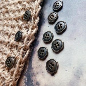 May include: Close-up of bronze buttons with four holes, scattered on a neutral background. Some buttons are attached to a section of woven, tan fabric. The buttons have a decorative, scalloped edge.