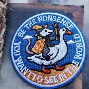 May include: A circular embroidered patch with a blue background featuring a white goose carrying a grey opossum wearing a yellow wizard hat and cape. The opossum holds a star-tipped wand. The text reads BE THE NONSENSE YOU WANT TO SEE IN THE WORLD.