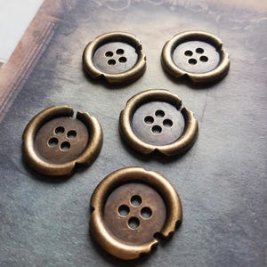 May include: Five antique brass buttons with four holes each. The buttons have a slightly distressed, weathered appearance, with a circular design and a raised outer rim. The buttons are arranged on a textured, muted background.