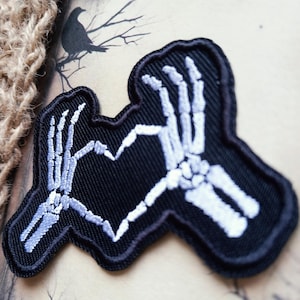 May include: A black embroidered patch featuring two white skeleton hands positioned to form a heart shape. The design is rendered in white thread against a solid black background with a thick black border outlining the shape of the hands.