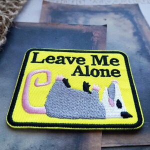 May include: A rectangular embroidered patch with a bright yellow background and a thick black border. The text Leave Me Alone is stitched in black letters above a grey and white opossum lying on its back with a pink tail and pink feet.