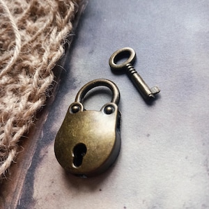 May include: A small, antique-style padlock and key set. The padlock is a rounded rectangle with a keyhole, and the key is a simple, slender design. Both pieces have a bronze finish, suggesting an aged or vintage aesthetic. The items are likely decorative or for small boxes.
