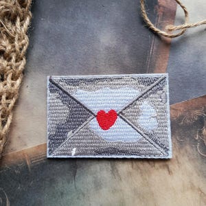 May include: A rectangular embroidered patch featuring a stylised envelope design. The envelope is rendered in shades of grey and white thread, with a prominent, solid red heart stitched at the centre where the envelope flap meets the body.