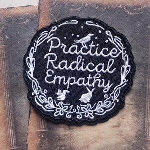 May include: Black embroidered patch with the words "Practice Radical Empathy" in white script. The patch has a scalloped edge and is decorated with white floral and animal illustrations, including a bird, squirrel, and rabbit.