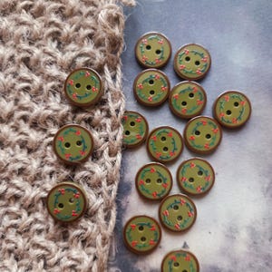 May include: A collection of round, olive green buttons, each featuring two central holes and a printed circular pattern of thin green vines with small red berries. The buttons are arranged in a cluster, with some resting on a textured brown fabric.