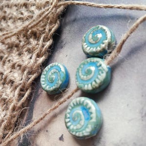 May include: Four ceramic beads with a spiral design in shades of blue and white are strung on natural twine. A piece of woven jute fabric is visible in the background, adding a rustic touch. The beads are round and have a glossy finish.