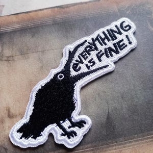 May include: A black crow patch with the text "EVERYTHING IS FINE!" in white. The crow is outlined in white, with a white border around the text. The patch is on a neutral background.