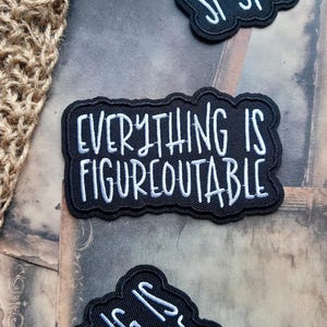 May include: A black embroidered patch with a scalloped edge featuring the white stitched text "EVERYTHING IS FIGUREOUTABLE" in a casual, hand-lettered style font. The patch has a dense, textured fabric base and a clean, finished border.