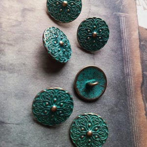 May include: Six antique-style buttons in teal and bronze. Each button has an ornate, floral design with a central raised dot. The buttons are arranged on a textured, neutral-toned surface, highlighting their intricate details and vintage aesthetic.