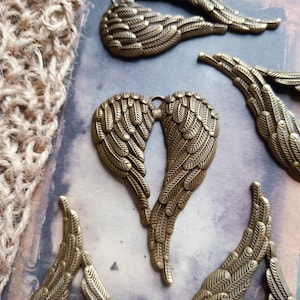 May include: A collection of antique bronze-coloured metal angel wing charms. Each charm features detailed, textured feather patterns. The wings are arranged in various positions, including a pair joined at the top to form a heart-like shape.