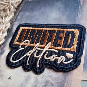 May include: A rectangular embroidered patch with a black border. The word LIMITED is stitched in metallic bronze thread inside a rectangular frame, while the word Edition is stitched in cream thread below it, finished with a small heart icon.