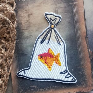 May include: An embroidered patch featuring a clear plastic bag tied at the top with a small yellow knot. Inside the bag is a small fish with a bright pink head and a vibrant orange body with yellow fins, rendered in detailed thread work.