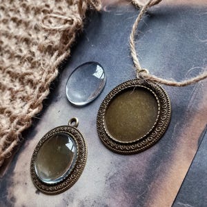 May include: A selection of jewellery-making components, featuring two bronze-toned pendant trays with ornate borders, a clear oval glass cabochon, and a small piece of woven fabric. The pendants are attached to natural twine.