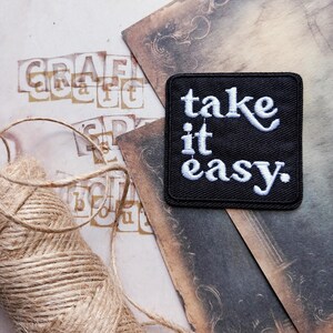 May include: A black square patch with white embroidered text reading "take it easy." The patch is set against a backdrop of aged paper and twine. The word "craft" is stamped in block letters.