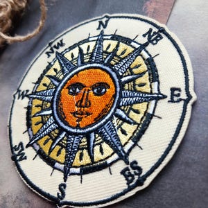 May include: A circular embroidered patch featuring a central orange sun face with human-like features, surrounded by black and yellow rays. The design includes a compass rose with black cardinal direction letters N, NE, E, SE, S, SW, W, and NW.