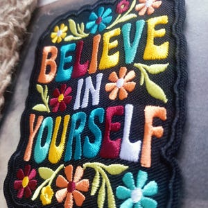 May include: A black embroidered patch with the words "BELIEVE IN YOURSELF" in vibrant colours. The patch is decorated with floral designs in shades of yellow, orange, red, and blue, and has a black border.