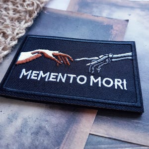 May include: A rectangular black embroidered patch featuring a white and brown human hand reaching towards a grey skeletal hand. The text MEMENTO MORI is embroidered in white capital letters below the hands, with a black border surrounding the design.