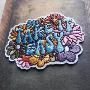 May include: Embroidered patch featuring the phrase "TAKE IT EASY" in light blue, set against a backdrop of colourful flowers in pink, purple, orange, and yellow. The patch has a white border and is designed to be sewn or ironed onto fabric.