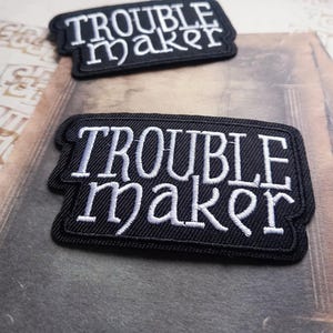 May include: Two black rectangular patches with the words "TROUBLE MAKER" embroidered in white. The patches are stacked, one above the other. The patches have a black border and are on a textured surface.