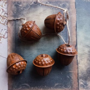May include: Five acorn ornaments in shades of brown, each suspended by twine. The acorns have textured caps and detailed grooves. The ornaments are arranged on a vintage-style background, evoking a rustic or autumnal aesthetic. The acorns are of varying sizes.
