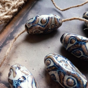 May include: Close-up of several oblong beads with a brown, white, and blue pattern. The beads are strung on a natural twine. The beads have a glossy finish and a unique, organic design. The background is a neutral, textured surface.