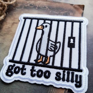 May include: A square embroidered patch featuring a white goose with an orange beak and feet standing behind black prison bars. A black lock is visible on the bars. The text "got too silly" is embroidered in black letters at the bottom of the patch.