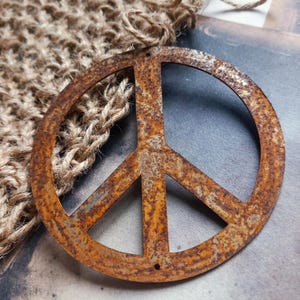 May include: A rusty metal peace sign ornament. The peace symbol is circular with three downward-pointing lines. The ornament has a distressed, aged appearance. A woven jute fabric and twine are in the background.