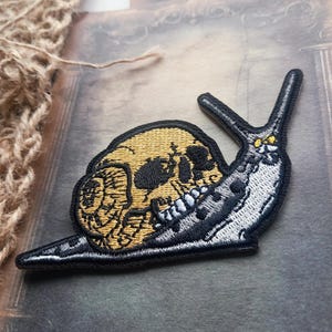 May include: An embroidered patch featuring a snail with a yellow human skull as its shell. The snail body is light grey with black spots and a black outline. The design includes two long antennae and a detailed skull with dark eye sockets and teeth.