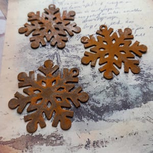 May include: Four rusty metal snowflake decorations with detailed cut-out patterns. Each snowflake has a small hole for hanging. The snowflakes are a warm, reddish-brown colour, suggesting an aged or weathered appearance. They are arranged on a vintage paper background.