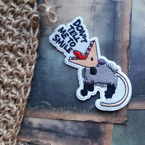 May include: An embroidered patch featuring a grey opossum with black limbs and a long tail, depicted with an open mouth showing sharp teeth and a red tongue. Above the opossum, the text reads DON'T TELL ME TO SMILE in black letters.