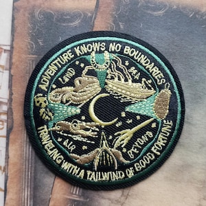 May include: Circular embroidered patch with the text "ADVENTURE KNOWS NO BOUNDARIES" in white. The patch features illustrations of a ship, hot air balloon, and other travel-themed elements in green and gold against a black background.
