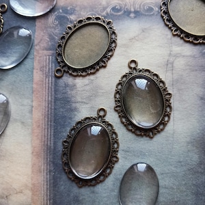 May include: A collection of antique bronze-toned oval pendant trays and clear glass cabochons. The pendant trays feature decorative, filigree-style frames with a loop for a chain. The cabochons are oval and slightly domed in shape.