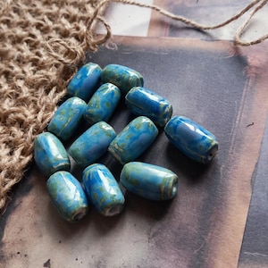 May include: A selection of blue and green ceramic beads. The beads are barrel-shaped with a glossy finish. The colour scheme features turquoise and teal hues, with a marbled pattern. The beads are arranged on a textured surface.