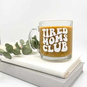 Op de afbeelding: Een heldere glazen mok met een bruine vloeistof erin. De mok heeft witte tekst die "TIRED MOMS CLUB" luidt in een retro-lettertype.