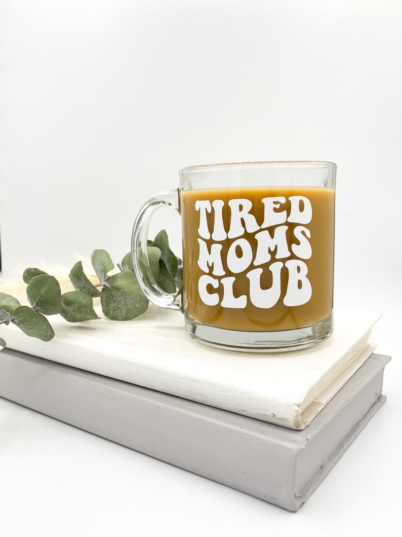 Op de afbeelding: Een heldere glazen mok met een bruine vloeistof erin. De mok heeft witte tekst die "TIRED MOMS CLUB" luidt in een retro-lettertype.