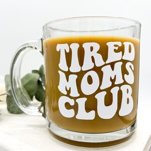 Op de afbeelding: Een heldere glazen koffiemok met een wit ontwerp waarop in een retro-lettertype "TIRED MOMS CLUB" staat. De mok is gevuld met koffie.