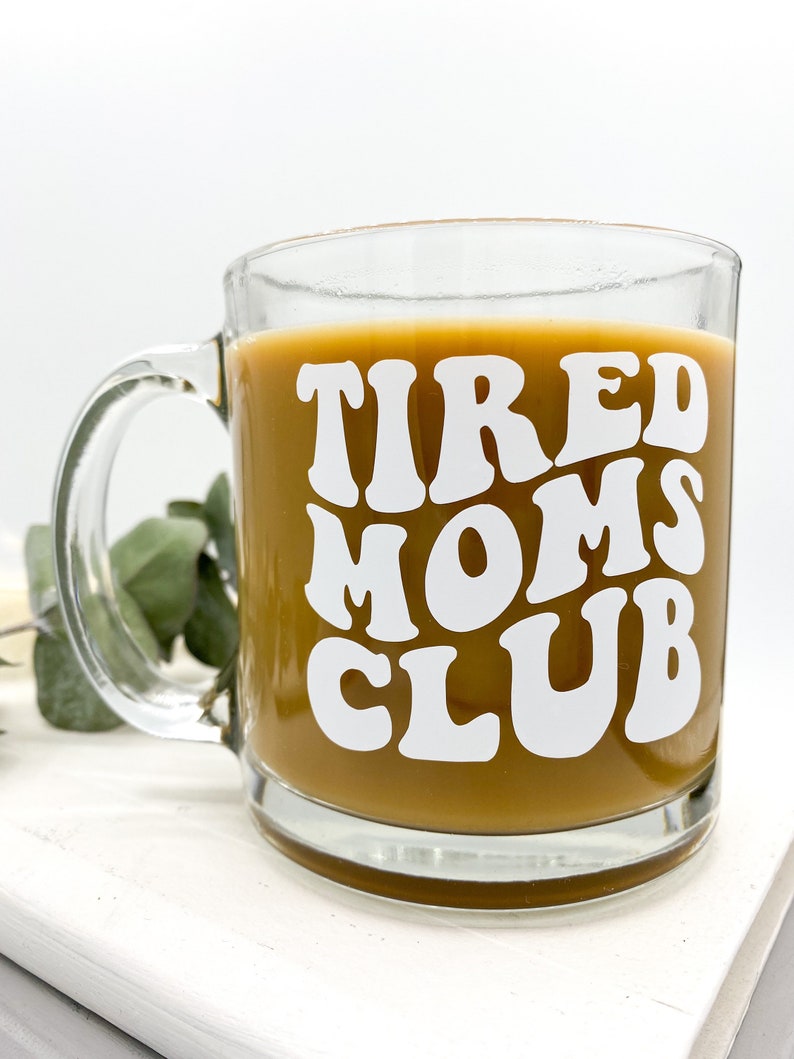 Op de afbeelding: Een heldere glazen koffiemok met een wit ontwerp waarop in een retro-lettertype "TIRED MOMS CLUB" staat. De mok is gevuld met koffie.