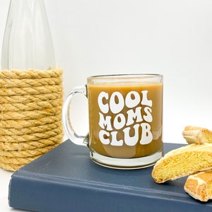 May include: A glass coffee mug with a white design that says "Cool Moms Club" in a retro font. The mug is filled with coffee and is sitting on a blue book with two biscotti cookies next to it.