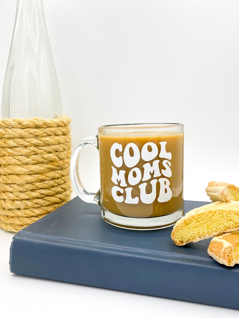 May include: A glass coffee mug with a white design that says "Cool Moms Club" in a retro font. The mug is filled with coffee and is sitting on a blue book with two biscotti cookies next to it.
