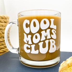 May include: A clear glass coffee mug with a white design that says "Cool Moms Club" in a retro font. The mug is filled with coffee and is sitting on a blue surface.