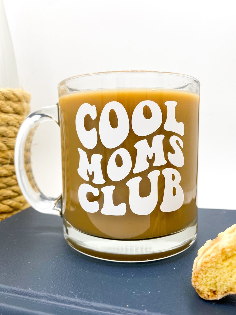 May include: A clear glass coffee mug with a white design that says "Cool Moms Club" in a retro font. The mug is filled with coffee and is sitting on a blue surface.