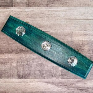 May include: A teal-painted wooden wall hook with three clear, faceted crystal knobs. The rectangular hook has a distressed finish, and small brass-colored hardware is visible on the back for hanging. The knobs are evenly spaced.
