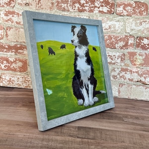 May include: A framed painting of a black and white border collie dog standing in a field with other black sheep. The dog is looking to the left of the frame. The background is a green field with a blue sky.
