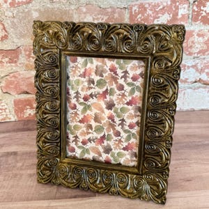 May include: Ornate gold-colored picture frame with a detailed, swirling design. The frame holds a print with a pattern of autumn leaves in shades of red, green, and brown. The frame is rectangular and stands upright on a wooden surface.