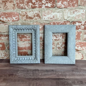 May include: Two gray picture frames with ornate designs. The frame on the left has a scalloped design and the frame on the right has a floral design.