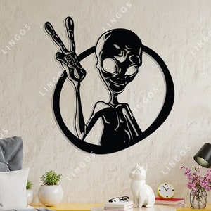 May include: Black metal wall art depicting an alien figure giving the peace sign, framed by a circle. The alien has large eyes and a smiling expression. The text "DXF / CDR / Ai / SVG / PDFS" is at the bottom.