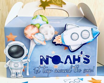 May include: A blue and white favor box with a space theme. The box features an astronaut, a rocket, planets, and stars. The text on the box reads "Noah's 1st trip around the sun!"