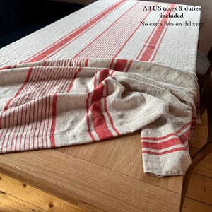 Peut inclure: Une nappe en lin avec un motif à rayures rouges et blanches. La nappe est drapée sur une table en bois, avec le tissu plié par endroits. Le texte "All US taxes & duties included" est visible en haut à droite.
